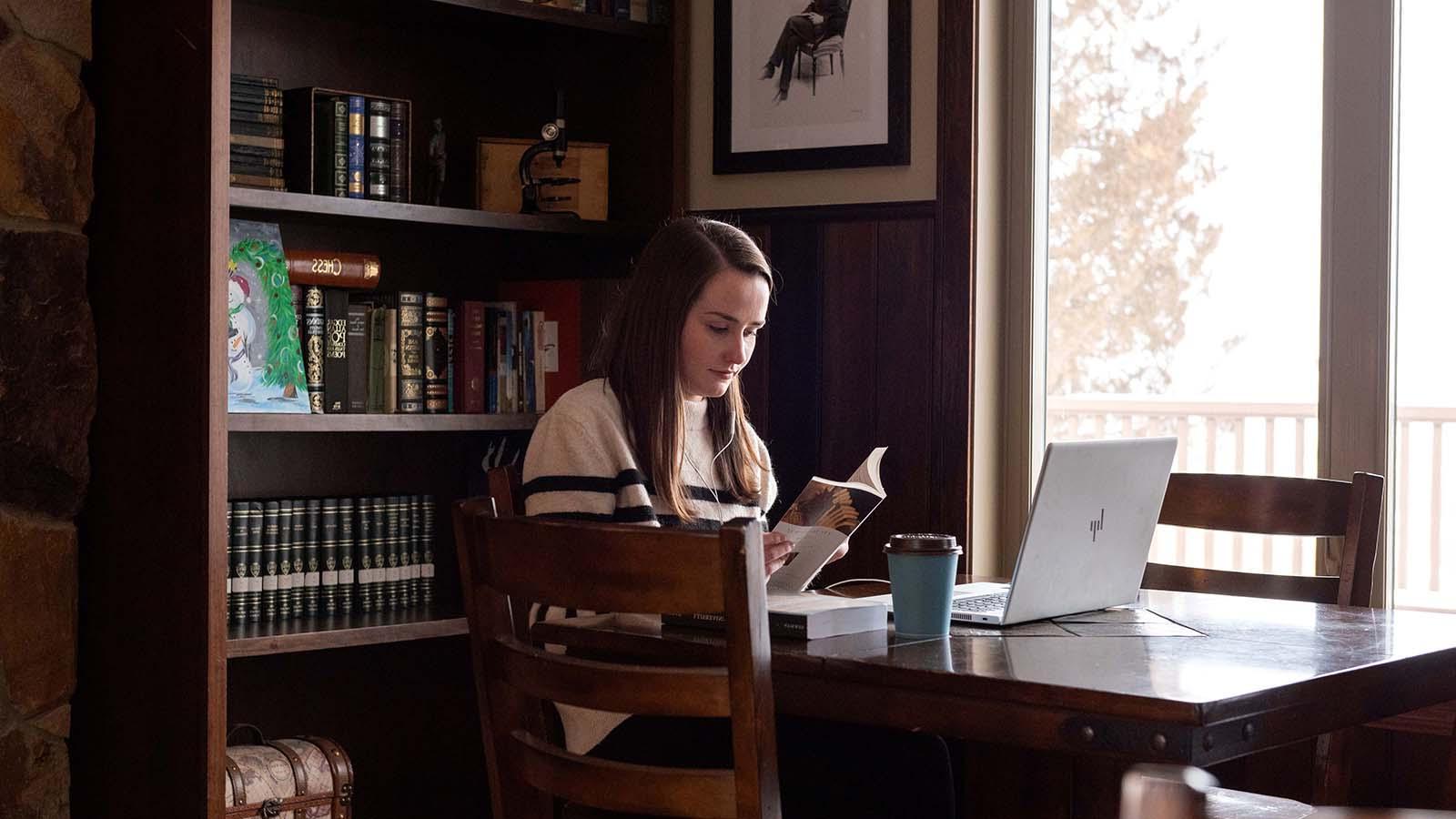 English student reading by window with coffee and laptop on table